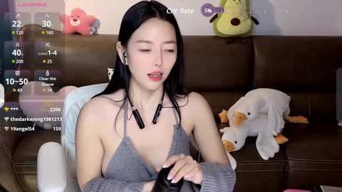 Media: Video of an Asian woman with long black hair, wearing a gray halter top, sitting on a brown couch, surrounded by plush toys, watching a video.