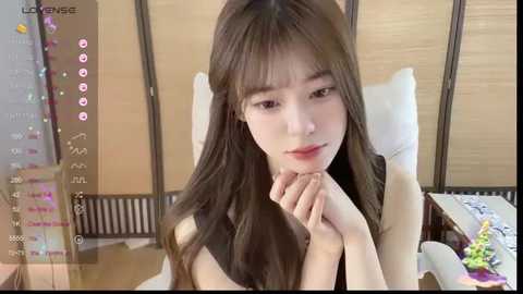 Media: A video of a young East Asian woman with long, straight brown hair, fair skin, and a petite frame, sitting indoors with a neutral expression. She wears a sleeveless black top.