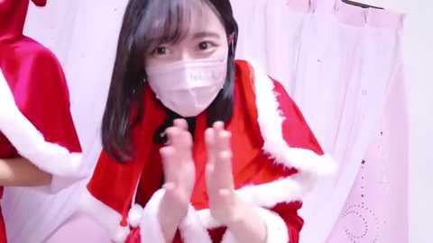 Media: A video of an Asian woman with long black hair, wearing a white face mask, red Santa dress, and white gloves, standing in front of white curtains with lace patterns.