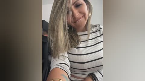 Media: Video of a smiling, fair-skinned woman with shoulder-length, straight, blonde hair, wearing a black and white striped long-sleeve top. She's holding a smartphone, revealing a colorful tattoo on her left arm.