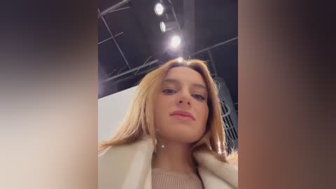 Media: Video of a young woman with fair skin and long, straight blonde hair, wearing a beige coat and a cream sweater, in a modern, dimly lit room with industrial ceiling lights.