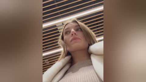 Media: Video of a blonde woman with fair skin, wearing a beige ribbed sweater, standing against a modern, wooden slatted ceiling. Her expression is neutral, eyes slightly downcast.