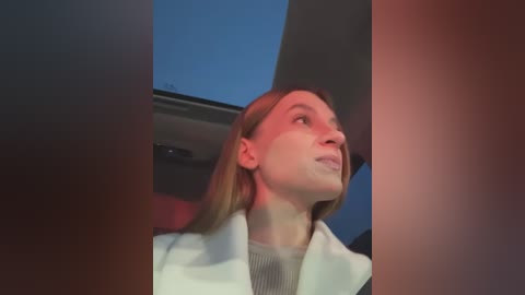 Media: Video of a blonde woman with fair skin, wearing a white coat, gazing upwards in a dimly lit room with a blue sky visible through a window.