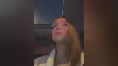 Media: A blurry video of a blonde woman with fair skin, wearing a white robe, sitting in a dimly lit room with a window showing a dark, possibly nighttime sky.