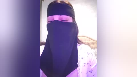 Media: Video of a person wearing a black niqab with a slit for the eyes, standing in a room with a white bedspread and visible headboard.