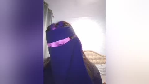 Media: A video captures a person in a black burqa, with pink headscarf, standing in a dimly lit bedroom with white walls and a bed with a wooden headboard.