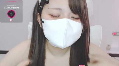 Media: Video of a young Asian woman with long, straight black hair, wearing a white surgical mask, sitting in a white chair, with a pink and white background.