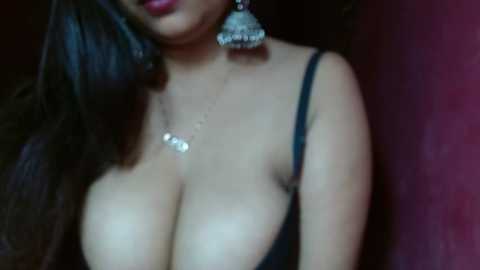 Media: Video of a woman with medium brown skin and long black hair, wearing a black spaghetti-strap top that reveals ample cleavage, adorned with large, ornate earrings.