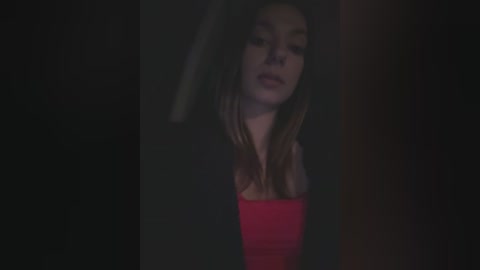 Media: A dimly-lit video featuring a young woman with long, straight brown hair, wearing a red top, partially obscured by a dark background.