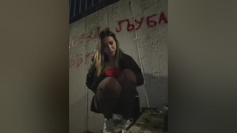 Media: Video of a young woman with long, blonde hair, squatting in a dimly lit, graffiti-covered alley. She wears a red top and dark pants, looking distressed.