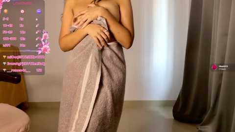 Media: Video of a nude woman with medium breasts, covered by a gray towel, standing in a bathroom with a flower-themed digital screen and a wooden chair.