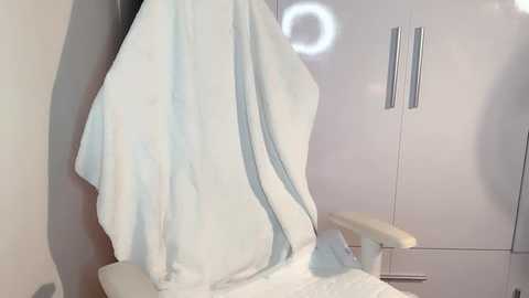 Media: Video of a white recliner chair with a blue and white blanket draped over it, against a white wall with silver handles and a circular light.