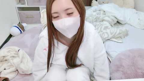 Media: Video of a young Asian girl with long brown hair, wearing a white face mask, sitting on a bed in a messy, pink-themed room with stuffed animals.