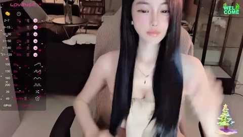 Media: Video of a topless East Asian woman with long black hair, pale skin, and a slender physique, sitting in a cozy living room with a Christmas tree, a fireplace, and a bookshelf in the background.