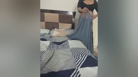 Media: Video of a woman in a black top and blue jeans, standing by a bed with a patterned quilt, wearing a blue blanket over her head, in a dimly lit room.