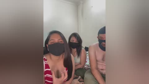 Media: Video of three people in masks, two women and one man, sitting on a bed in a dimly lit room. The women have medium skin tones and the man has a dark complexion.