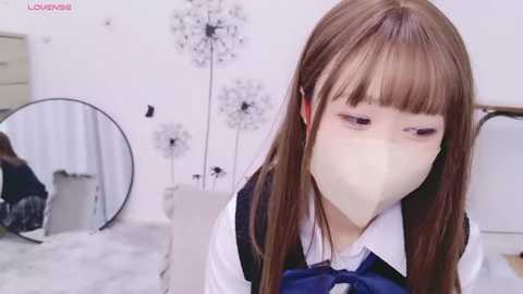 Media: Video of a young Asian woman with long brown hair and a white face mask, wearing a blue and white school uniform, standing in a minimalist, white-walled room with dandelion art and a mirror.