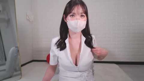 Media: Video of an Asian woman with long black hair, wearing a white short-sleeved blouse with a deep V-neck, white surgical mask, and white gloves, in a sterile room with white tiled walls and a glass shower door.