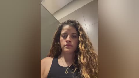 Media: Video of a young woman with long, wavy brown hair, fair skin, wearing a black tank top, standing in a dimly lit hallway.