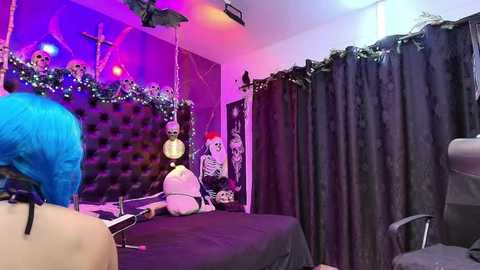 Media: Video of a dark, gothic bedroom with a tufted black headboard, purple and pink lighting, a skeleton figure on a bed, and a blue-haired person seated.