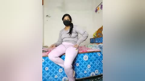 Media: Video of a woman with medium skin tone and black hair, wearing a black face mask, gray long-sleeve top, and pink pants, seated on a blue-patterned bed in a simple, cluttered room.