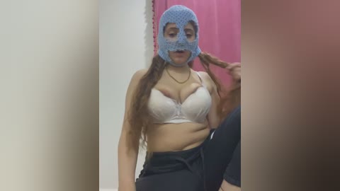 Media: Video of a woman with long brown hair, wearing a blue polka-dot mask, white lace bra, and black pants, sitting in a bathroom with pink and beige walls.