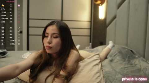 Media: A video of an East Asian woman with long brown hair, lying on a beige bed, wearing a white shirt, in a dimly lit bedroom with minimalist decor.