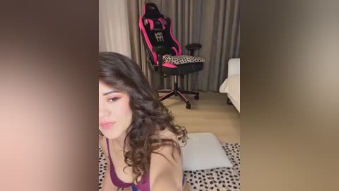 Media: Video of a young woman with long curly brown hair, wearing a purple top, sitting on a leopard-print rug in a modern bedroom with beige curtains, a gaming chair, and a bed.