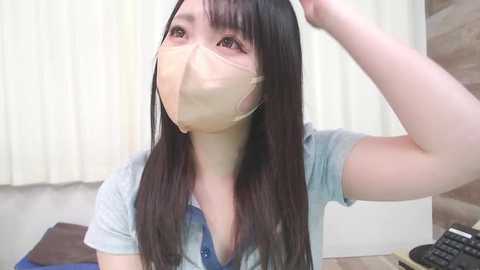 Media: A video of a young East Asian woman with long black hair, wearing a beige surgical mask, blue shirt, and light blue jeans, adjusting her hair. She's indoors with white curtains and a wooden wall in the background.