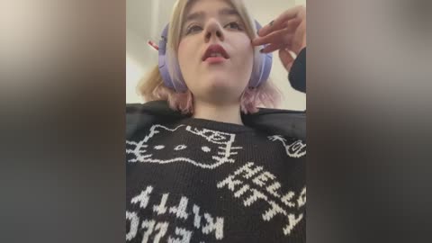 Media: Video of a young woman with short, blonde hair, wearing large purple headphones, and a black sweater with a white cat design. She has a neutral expression and is touching her ear.