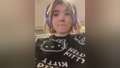 Media: Video of a young person with shoulder-length blonde hair, wearing purple headphones and a black sweater with a \"Hello Kitty\" design. Background shows a kitchen with cabinets.