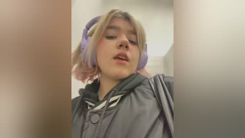 Media: Video of a young woman with light skin, shoulder-length blonde hair, wearing large purple headphones, and a grey raincoat, standing in a dimly lit indoor hallway.