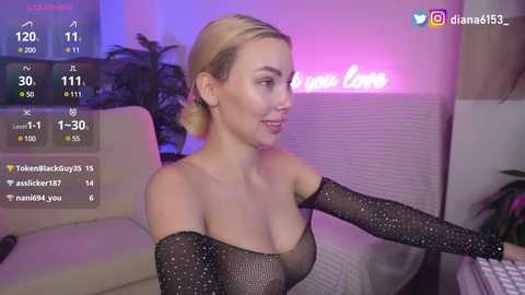 Media: Video of a blonde woman with light skin, wearing a black, glittery off-shoulder top, smiling and holding a controller, in a cozy living room with a neon sign and greenery.