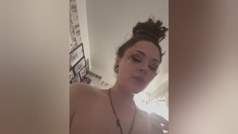 Media: A video of a topless young woman with fair skin, brown hair in a bun, wearing dramatic makeup, standing in a bathroom with a mirror reflecting her.