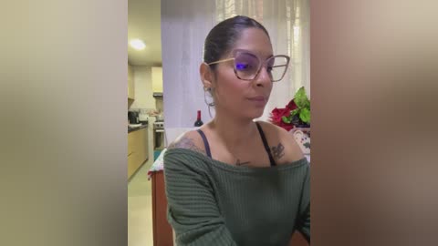 Media: A video of a Latina woman with a medium skin tone, dark hair, and glasses, wearing a green off-shoulder top, standing in a modern kitchen with white cabinets and a tiled backsplash.