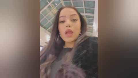 Media: A video of a young woman with long, straight brown hair, wearing a black fur coat and dark makeup, standing in a subway station with green-tiled walls and a glass ceiling.