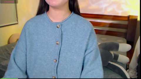 Media: A video of a young person with long dark hair, wearing a light blue, button-up sweater, standing in a bedroom with a wooden bed frame and gray bedding.