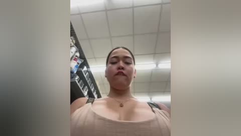 Media: Video of a young woman with medium skin tone, brown hair, wearing a beige tank top, taken from a low angle in a fluorescent-lit room with white ceiling tiles and a ladder.