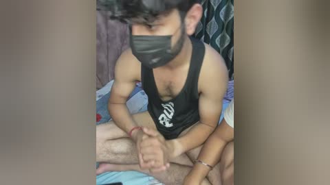 Media: Video of a South Asian man with short black hair, wearing a black mask and tank top, seated on a bed, hands tied behind his back.