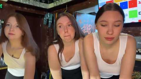 Media: Video of three young women in white tank tops, leaning forward, in a dimly-lit bar setting with wooden decor and a flat-screen TV.