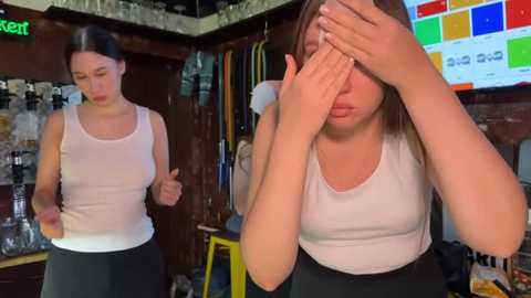 Media: Video of a distressed young woman with long brown hair, wearing a white tank top and black pants, covering her face in a cluttered, dimly lit room.