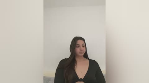 Media: Video of a young woman with long, dark hair, wearing a black top, standing in a minimalist white room with a simple bed.