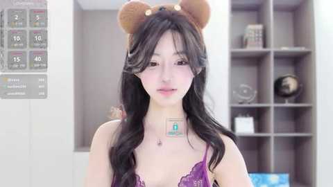 Media: Video of a young Asian woman with long black hair, wearing a teddy bear headband, purple lace bra, and a medical ID tag, in a modern room with a wall calendar and shelving unit.