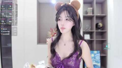 Media: A video of a fair-skinned East Asian woman with long black hair, wearing a purple lace bralette, brown bear ears headband, and sitting in a modern room with white walls and shelves.