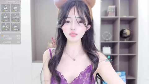 Media: A video of a young East Asian woman with long black hair, wearing a purple lace bra and a bear headband, standing in a modern, well-lit room with a temperature display and shelves.