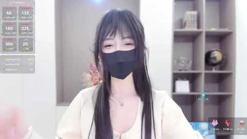 Media: Video of an Asian woman with long black hair and a black face mask, wearing a beige top, standing in a modern office with a monitor displaying weather data, and a bookshelf in the background.