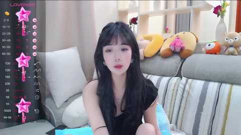 Media: A video of a young East Asian woman with long black hair, fair skin, and red lipstick, wearing a black top, sitting on a bed with stuffed toys and a window.