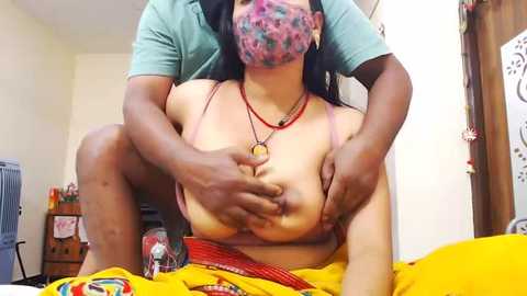 A video of a topless Black woman with medium-sized breasts, wearing a face mask, being held by a man in a teal shirt. Background shows a cluttered room with a yellow bedspread.