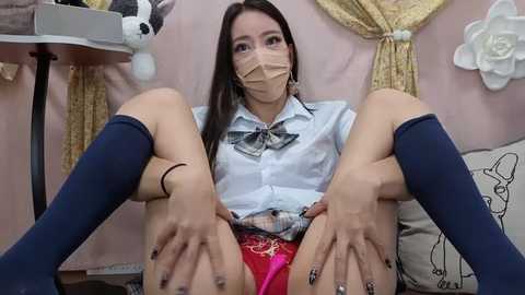 Media: Video of an Asian woman with long black hair, wearing a white blouse, red skirt, blue knee-high socks, and a surgical mask, sitting with legs spread, revealing red panties. Background features pastel-colored walls and decorative elements.