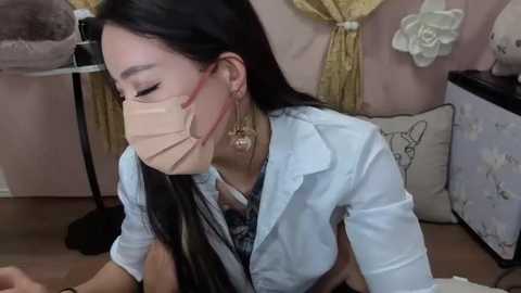 Media: Video of an Asian woman with long black hair, wearing a surgical mask and light blue shirt, kneeling in a room with beige walls, floral decor, and a plush toy.